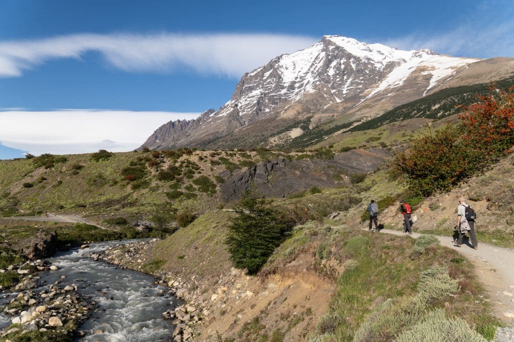 Hikers walking a path next to a stream leading up to a snowy mountain in Patagonia.