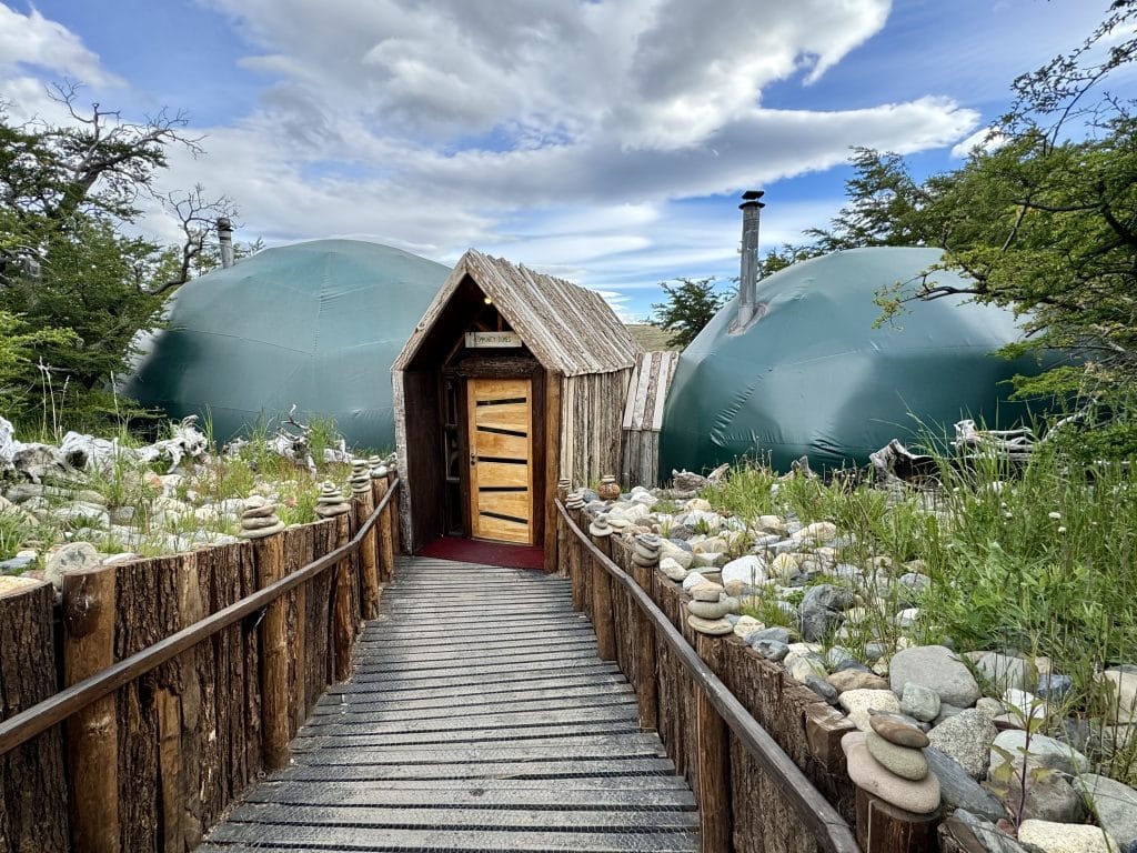 A little wooden house at the end of a wooden pathway, set between two green domes.