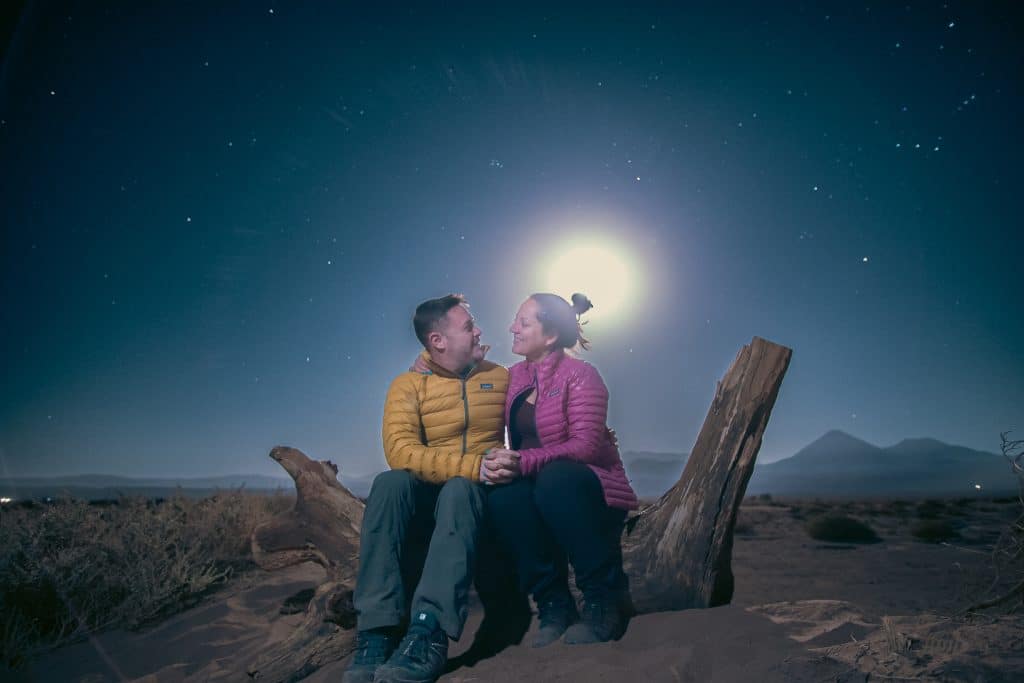 Kate and Charlie, in their pink and yellow Patagonia jackets, sitting on a log in the desert, underneath a night sky with a very bright full moon.