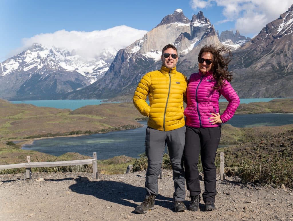 Charlie and Kate, in matching Patagonia coats (his yellow, hers hot pink), standing in front of a jagged gray-purple mountain topped with snow and bright turquoise lakes. Kate's long wavy brown hair is flailing in a million directions due to the wind.