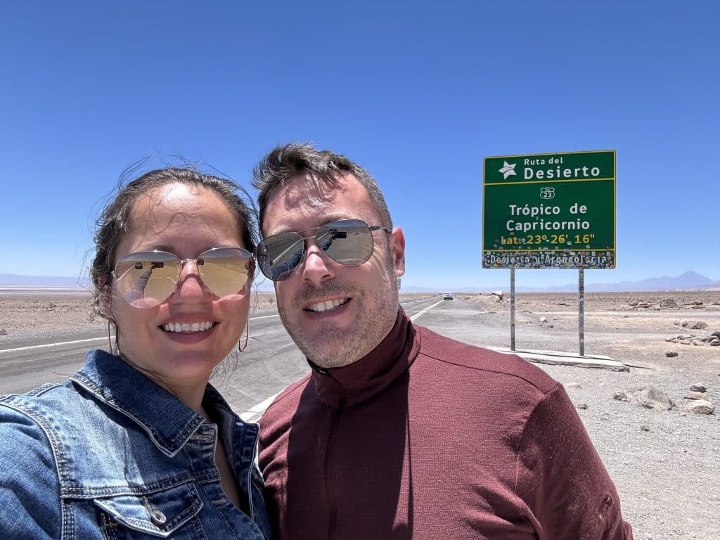 Kate and Charlie taking a smiling selfie in sunglasses in front of a road sign reading Tropico de Capricornia.