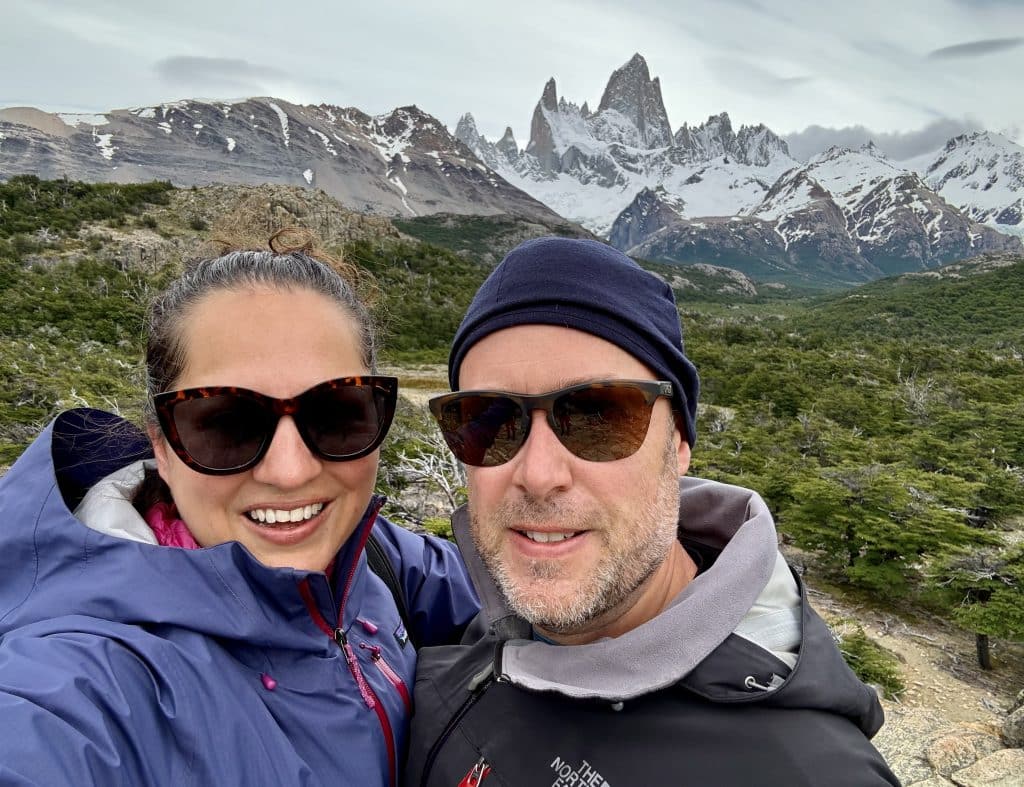 Kate and Charlie taking a smiling selfie in sunglasses and hiking gear, in front of the Mount Fitz Roy jagged mountain viewpoint.