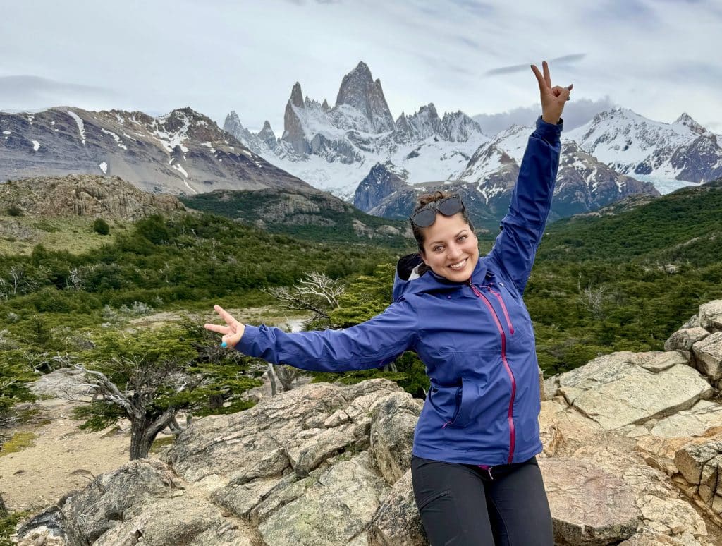 Kate posing with two peace signs in front of the Fitz Roy mountain range, jagged gray mountains surrounded by clouds.