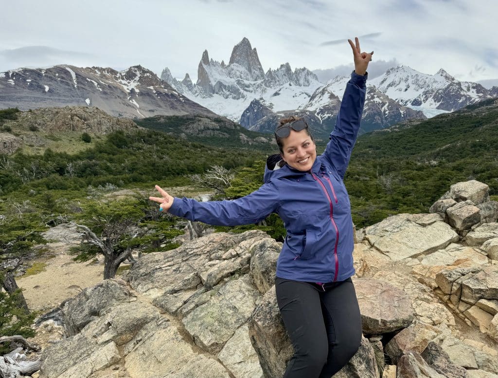 Kate posing with two peace signs in front of the Fitz Roy mountain range, jagged gray mountains surrounded by clouds.