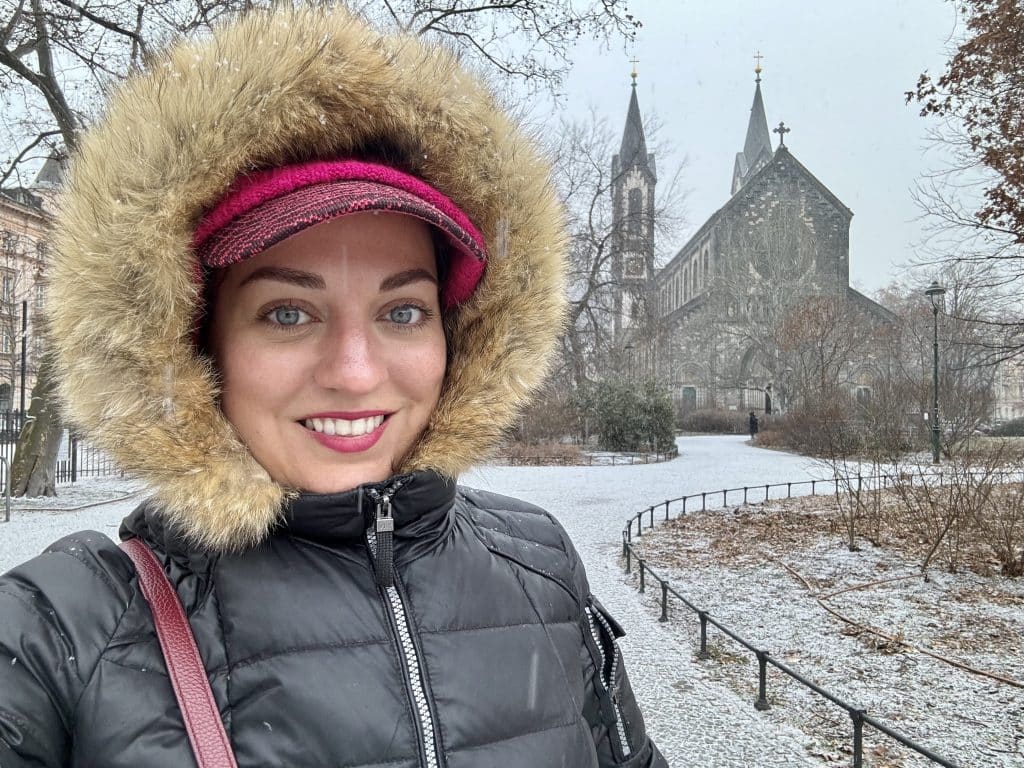 Kate prend un selfie souriant, vêtue d'un grand manteau d'hiver avec une capuche en fourrure. Elle est sur une place enneigée de Prague, une église gothique derrière elle.