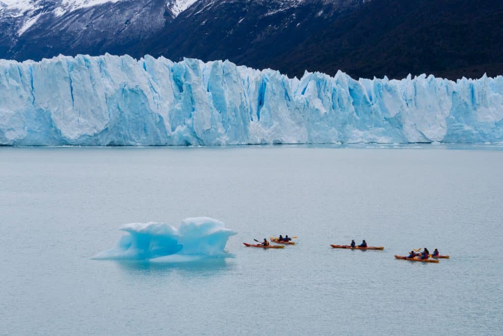 People in kayaks in a small lake in front of a massive blue and white glacier.