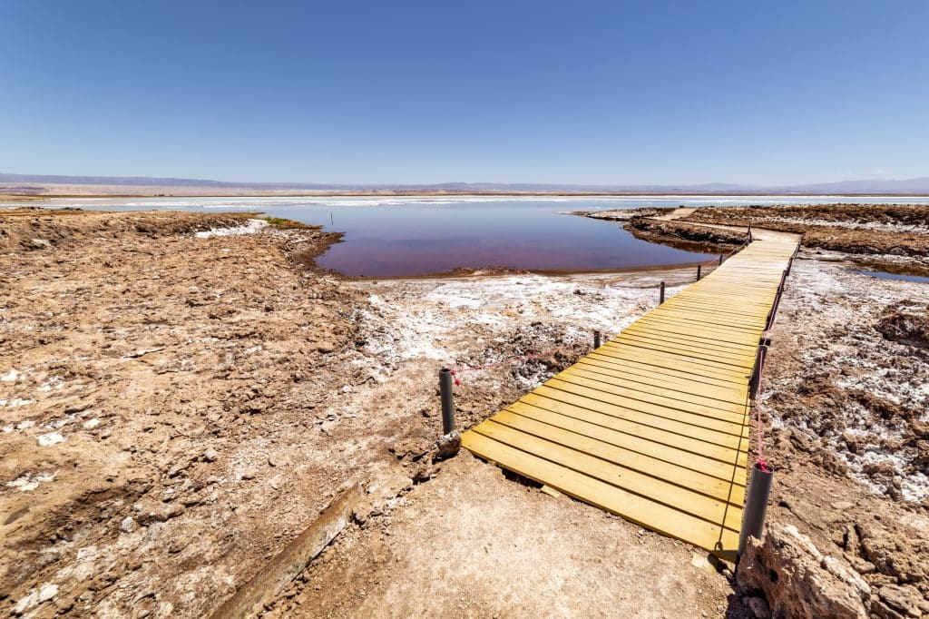 A yellow wooden platform leading over a salt-streaked sandy landscape and a blue still lagoon.