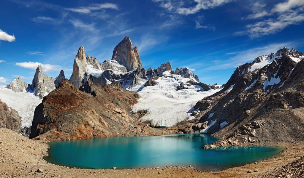An intensely teal lake surrounded by jagged mountains covered with snow underneath a blue sky.