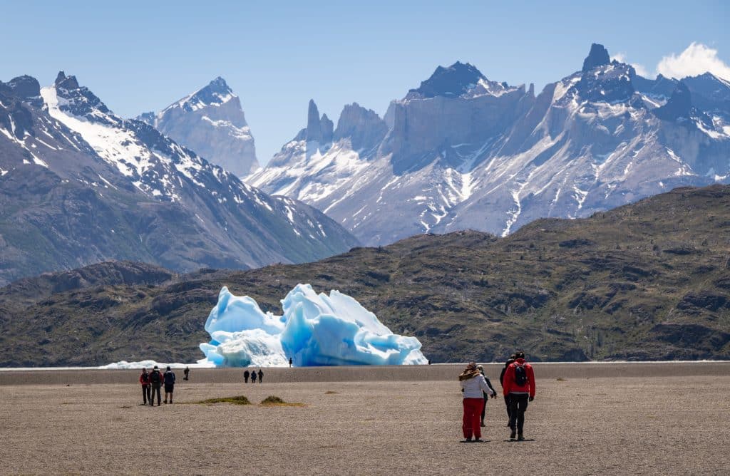 People walking on a gray sand beach, with a bright blue and white iceberg in the distance, floating on a lake. There are jagged purple-gray mountains in the distance.