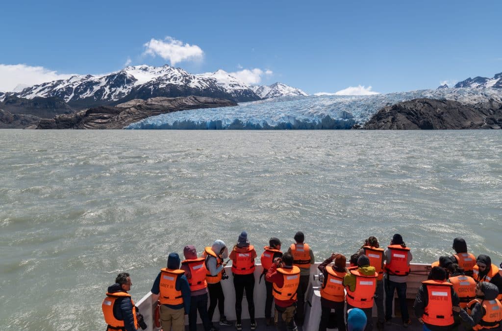 A group of people in orange life jackets standing on the edge of a boat, overlooking a steely gray lake and a blue and white glacier in the distance.
