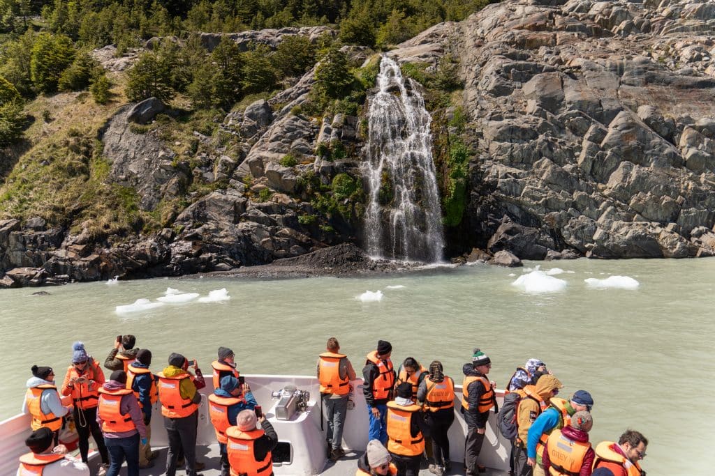 A group of people in orange lifejackets standing on the edge of a boat. In front of them is a waterfall softly falling from a rocky cliff, almost like a bridal veil, falling into a gray-green lake.