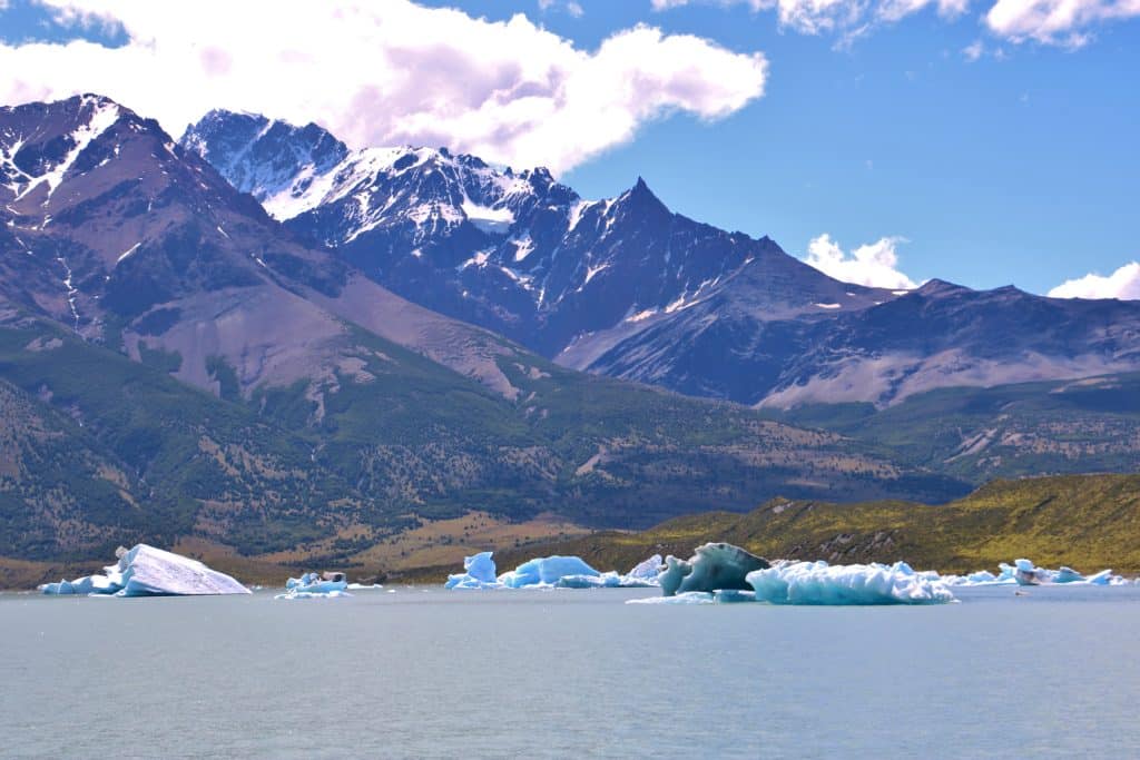 Icebergs floating on a still blue lake, a big purple mountain behind it.