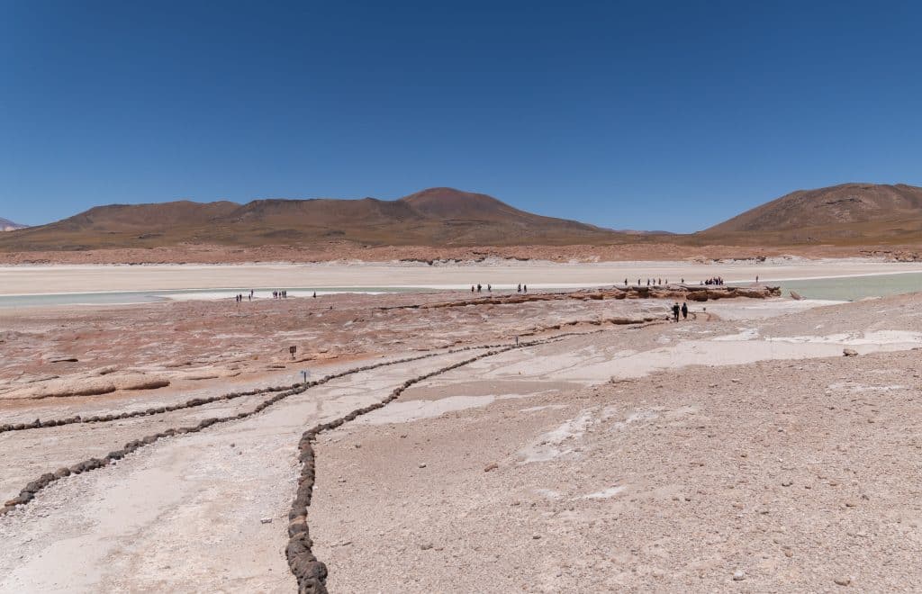 A pink desert landscape with red mountains and a pale blue-green lagoon with a trail marked with rocks on each side of it.