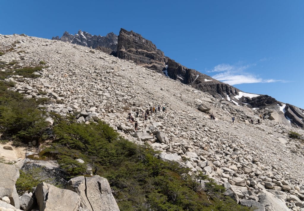 A mountainside with gray scraggling rocks, and teeny tiny hikers hiking among them.