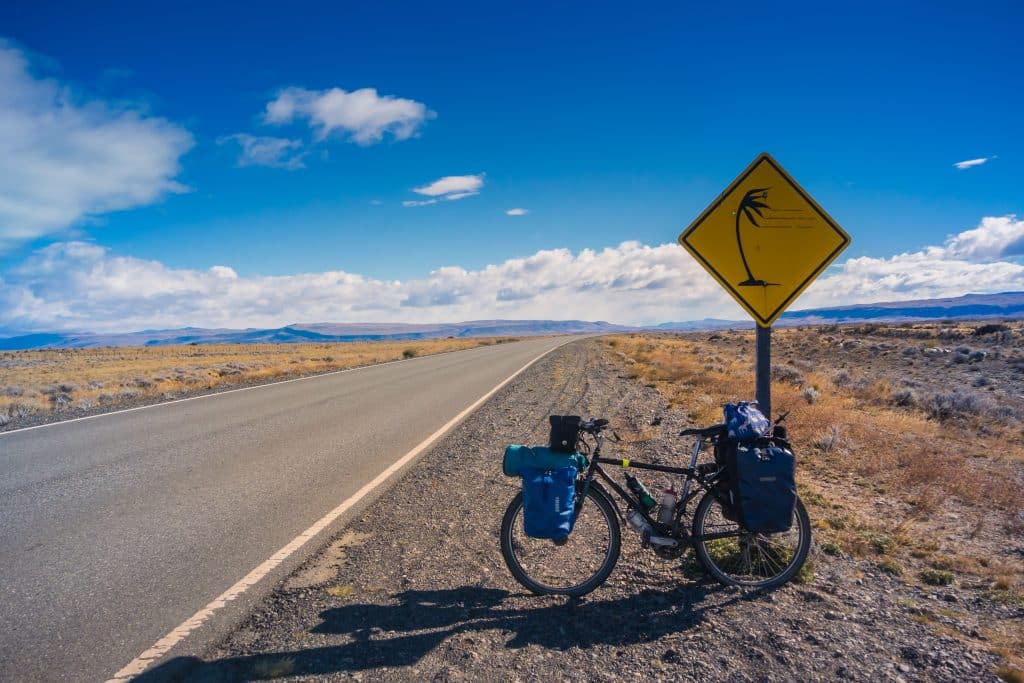 A bicycle parked by the side of the road in a flat part of Patagonia, a sign showing a tree being blown wildly to one side.