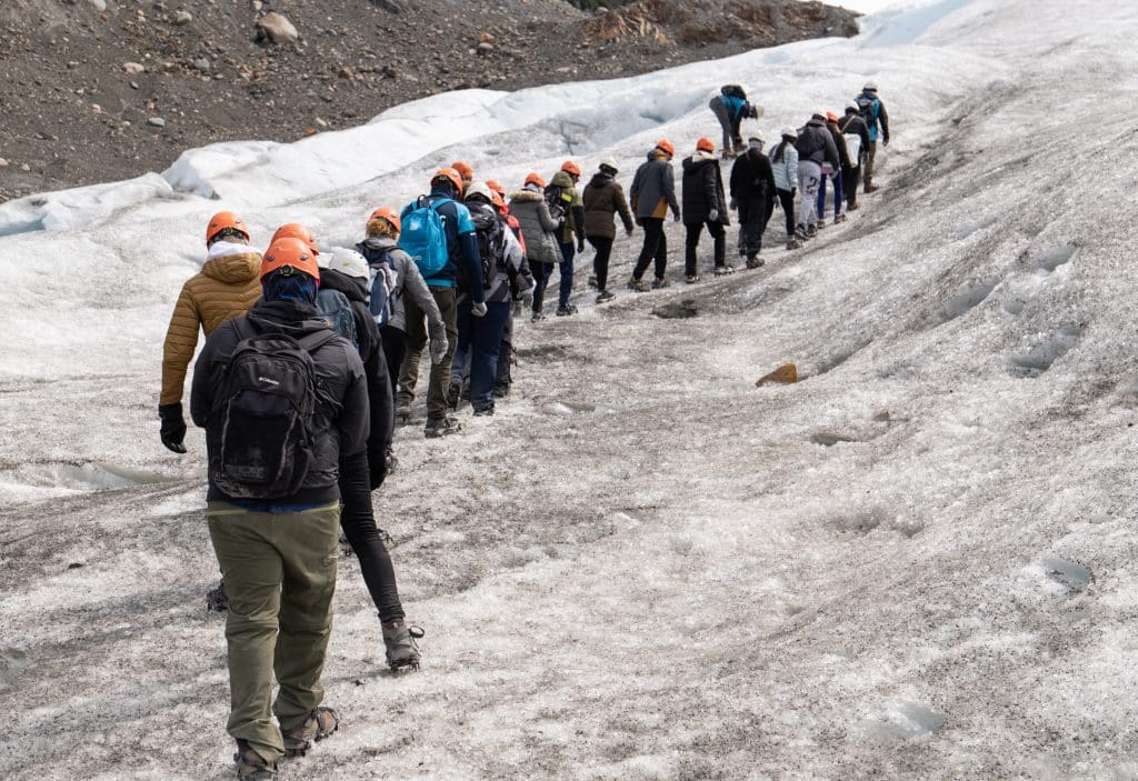 A line of about 20 people hiking on a glacier, everyone wearing helmets.