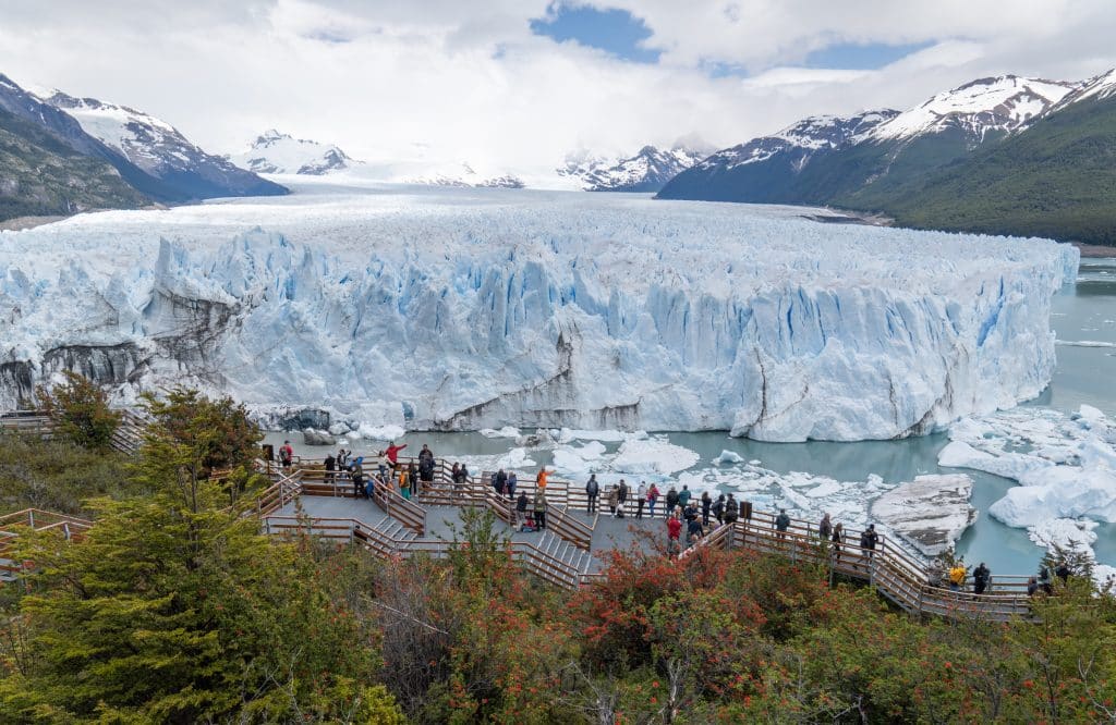 An enormous blue and white glacier surrounded by snow-capped mountains, and in the foreground, a walkway covered with dozens of people posing for photos.