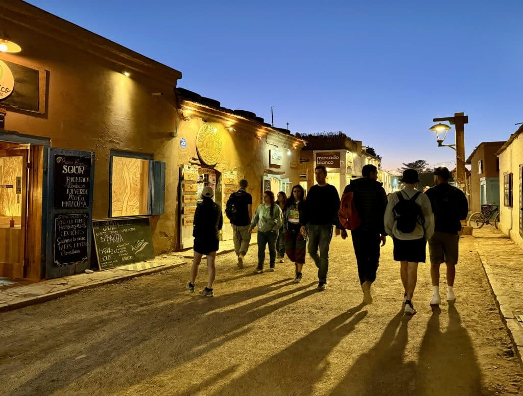 People walking down a dirt road in San Pedro de Atacama, just after sunset, the sky bright blue, and mud buildings on each side of the street.