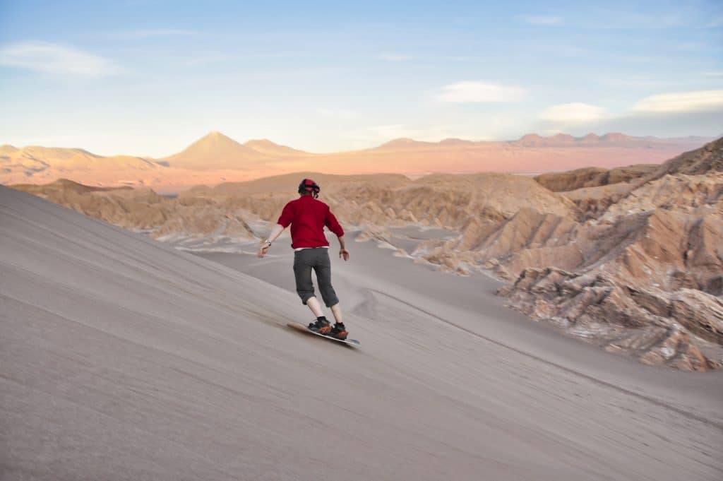 A man in a red shirt sand boarding down a dune in the atacama desert, pinkish mountains rising behind him.
