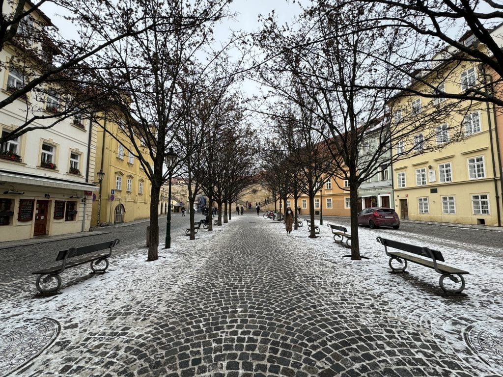 A cobblestone street in Prague, dusted with a layer of snow and surrounded by yellow buildings.