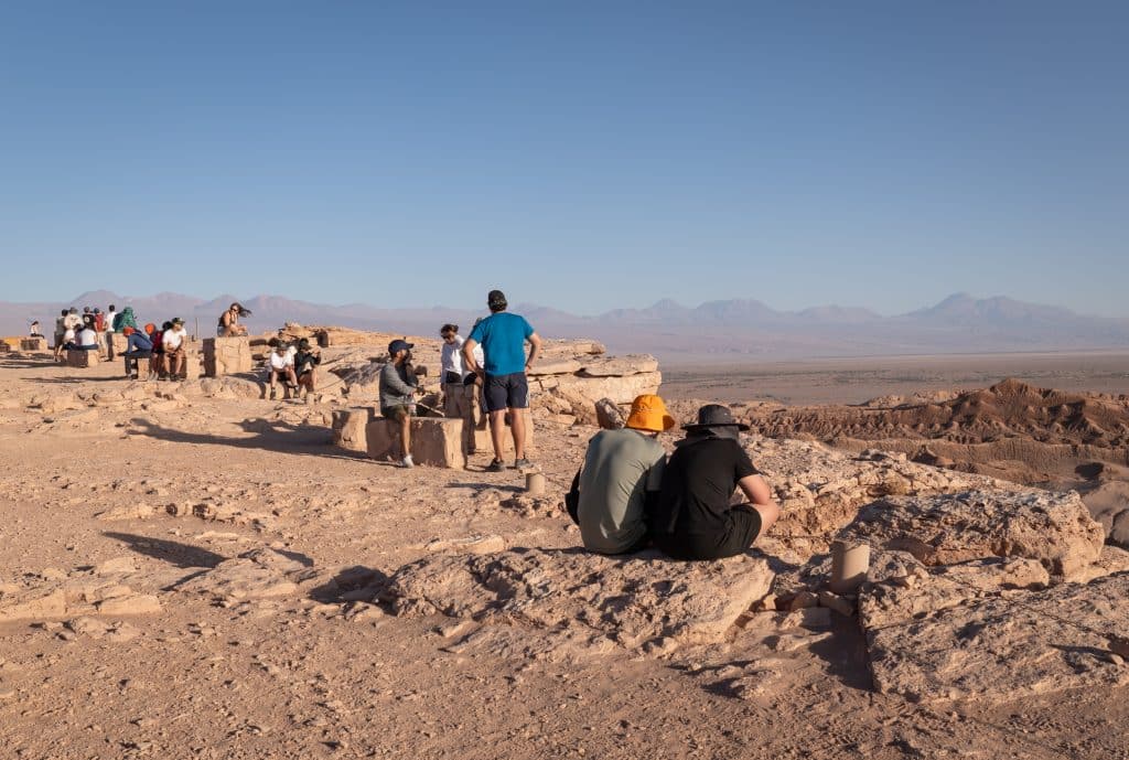 People hanging out on the edge of a rocky cliff in the desert, getting ready to watch the sunset.