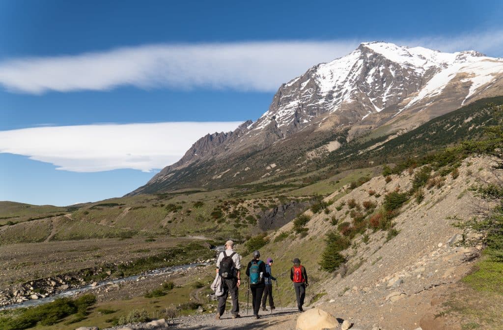 A few people hiking on a path leading up to a snow-covered mountain. The clouds are weird -- like two thick, solid lines.