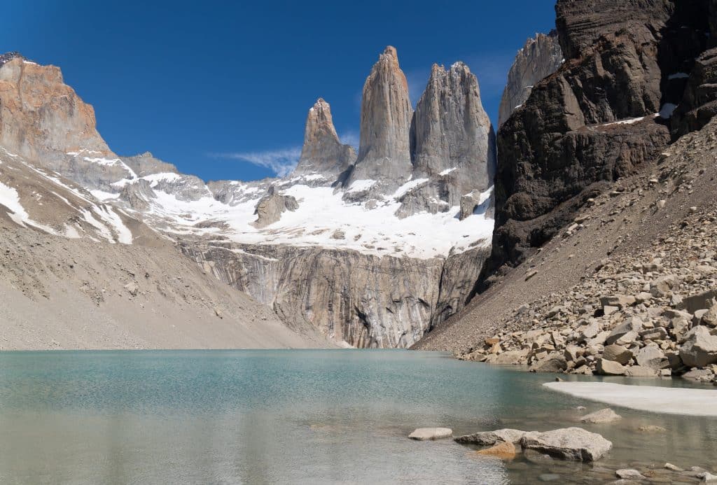 A placid clear turquoise lake surrounded by gray rocks, including three jagged towers behind it.