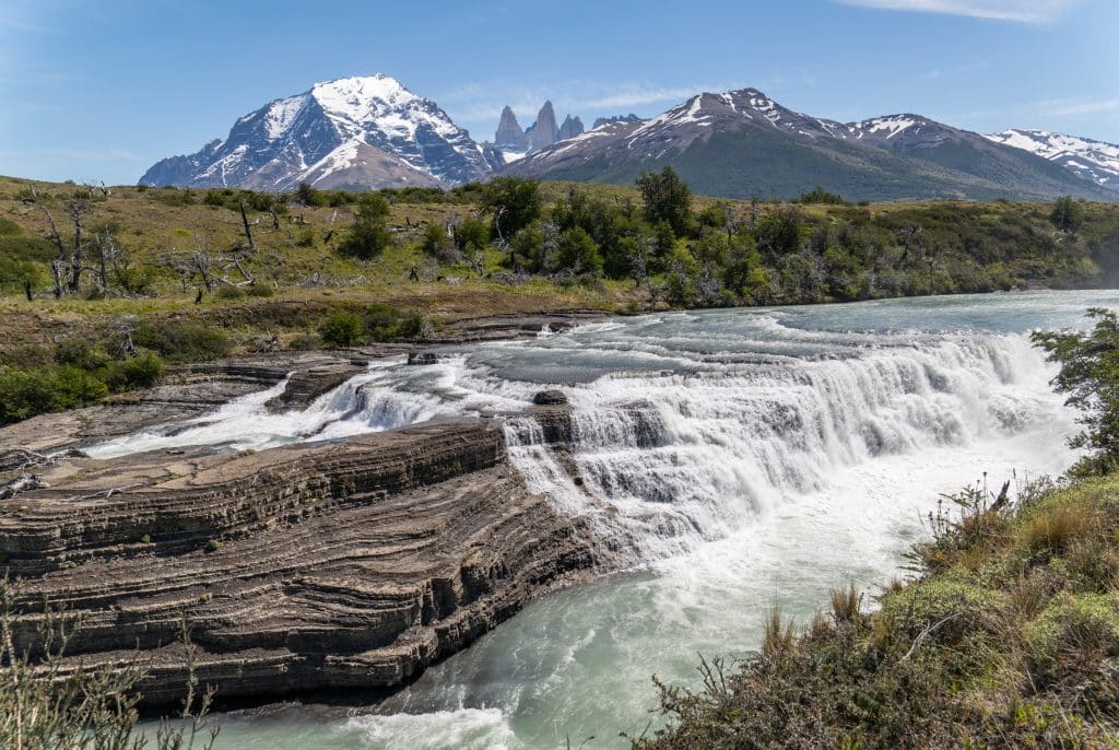 A large waterfall flowing over a huge rock, purple-gray mountains and the three jagged towers behind it.