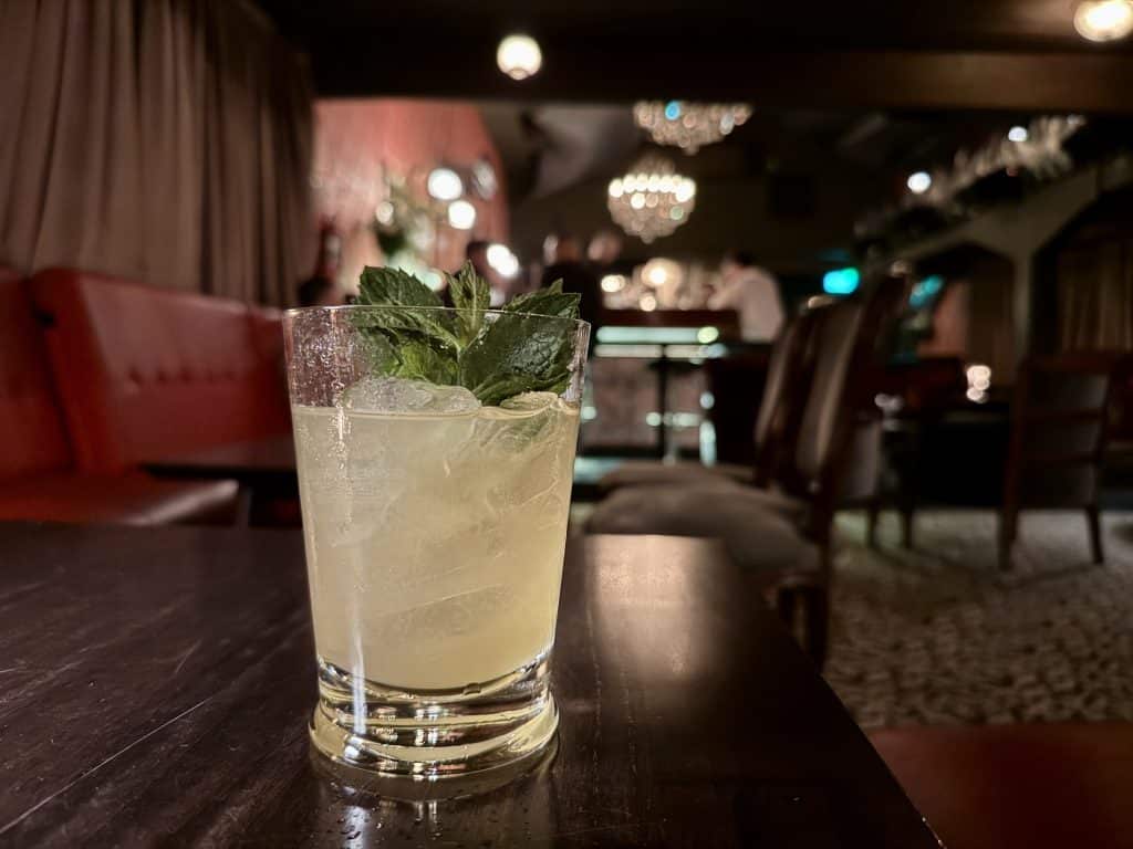 A pale yellow cocktail on a wooden table in a dark speakeasy-style bar.