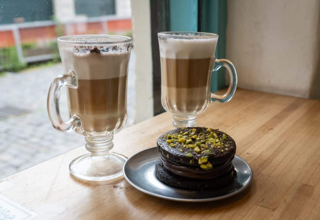 Two tall lattes in glasses with handles, next to an alfajor pastry covered in dark chocolate and pistachios.