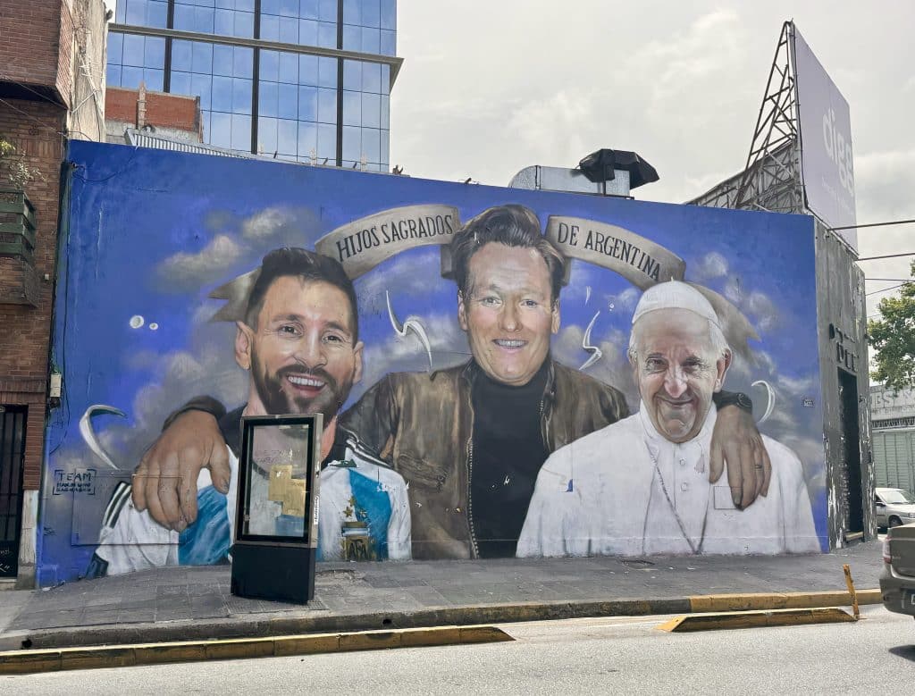 A mural of Conan O'Brien, Lionel Messi, and Pope Francis, with "sacred sons of Argentina" written in Spanish above them.