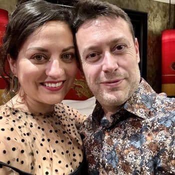 Kate and Charlie taking a smiling selfie. She wears a black polka dot sheer top over a black tank top; he wears an abstract blue and brown patterned shirt.