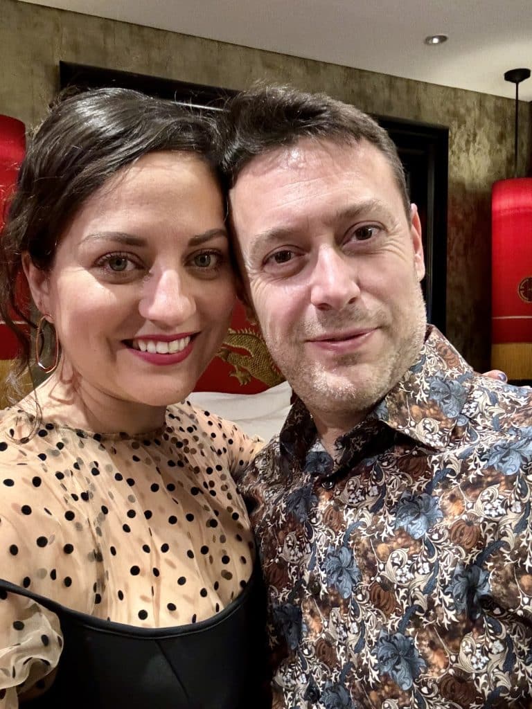 Kate and Charlie taking a smiling selfie. She wears a black polka dot sheer top over a black tank top; he wears an abstract blue and brown patterned shirt.