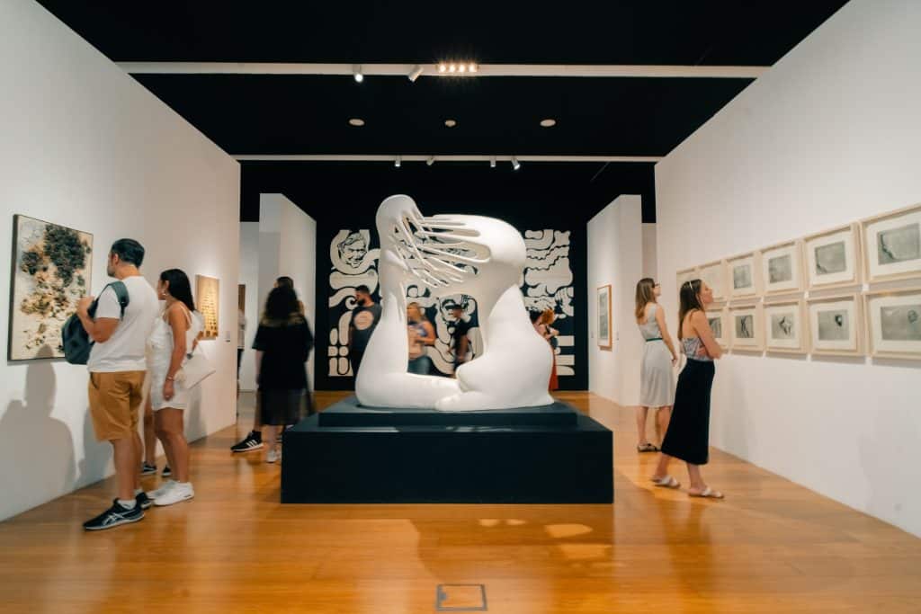 People walking around an art museum with a white abstract sculpture on the floor and paintings on the walls.