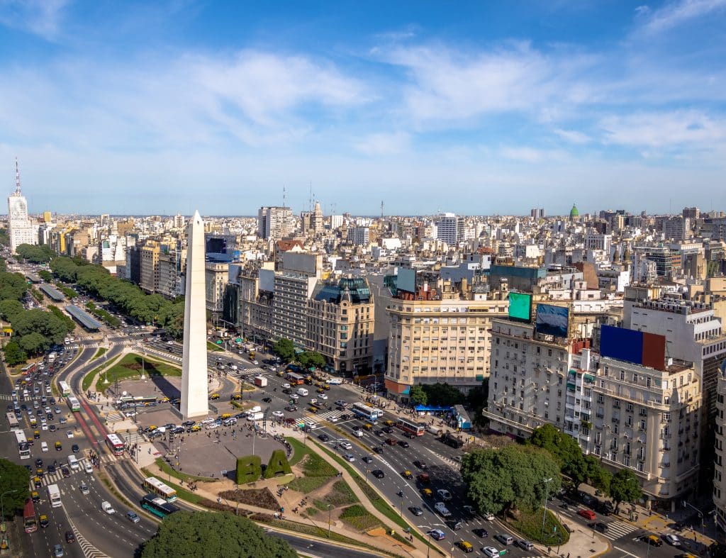 An aerial shot of Buenos Aires with a giant obelisk piercing the sky, lots of traffic on the streets, and lots of tall buildings.