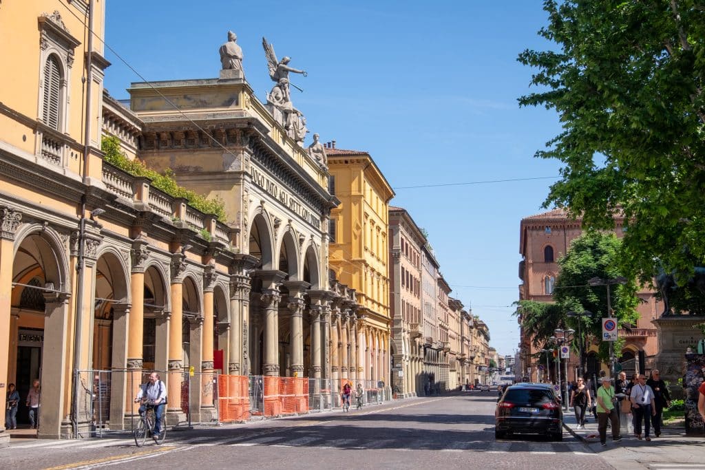 A city street in Bologna with lots of porticoes on one side of the street, and a single man riding a bicycle.