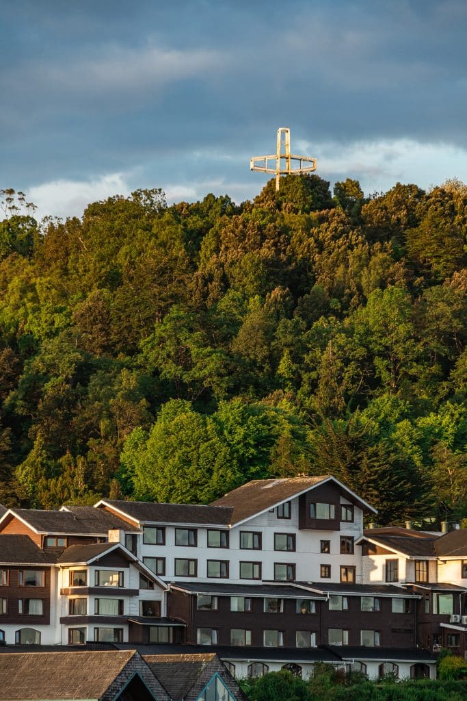 A house sitting at the bottom of a tree-covered hill topped with a cross.