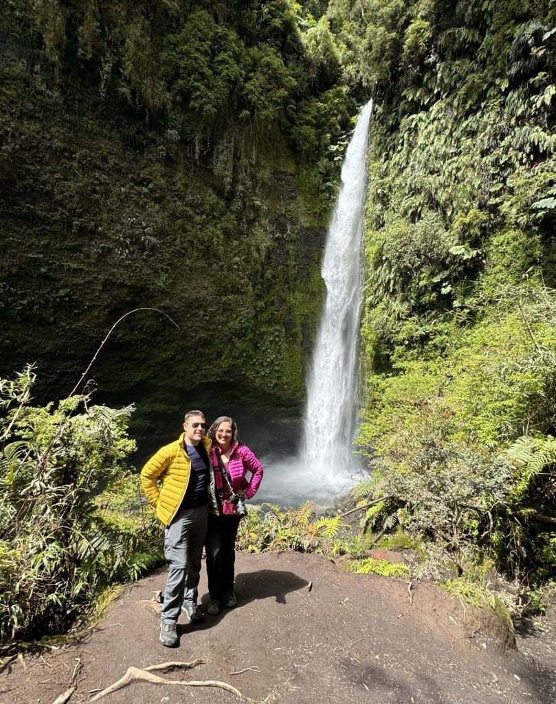 Kate and Charlie, in matching Patagonia jackets (his yellow, hers hot pink), standing next to a tall, skinny waterfall surrounded by mossy green cliffs.