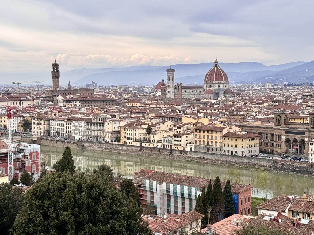 The city skyline of Florence with its Duomo, towers, and the green Arno River, underneath a cloudy sky.
