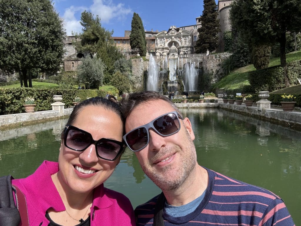 KAte and Charlie taking a smiling selfie in front of fountains at an Italian villa.