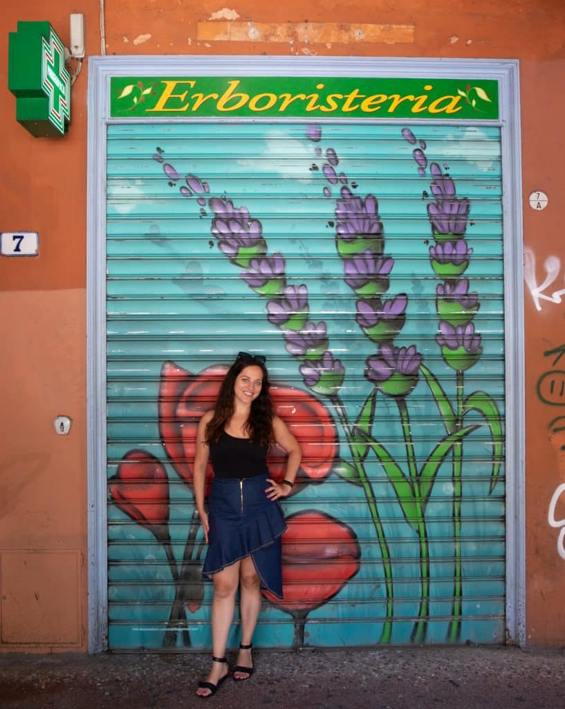 Kate standing in front of a graffitied wall in Bologna covered with purple and pink flowers.