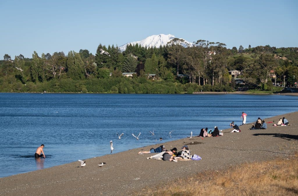 People sunbathing on a dark gray lake beach in front of a deep blue lake. One guy is in the lake up to his waist. There's a snowy mountain in the distance.