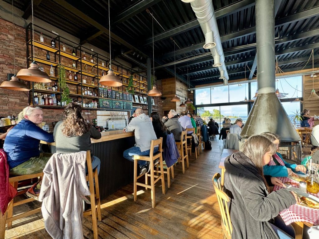 A restaurant with people sitting at a wooden bar, and big wood stoves reaching down from the ceiling.