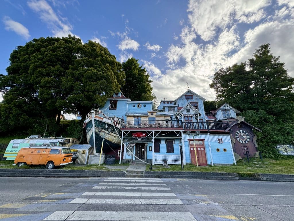 An museum building shaped like a house that has a giant cuckoo clock next to it, a boat built into the side of the house, and two VW vans parked next to it.