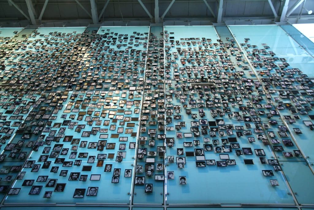A museum exhibit with hundreds of photos of people set on a blue glass wall.