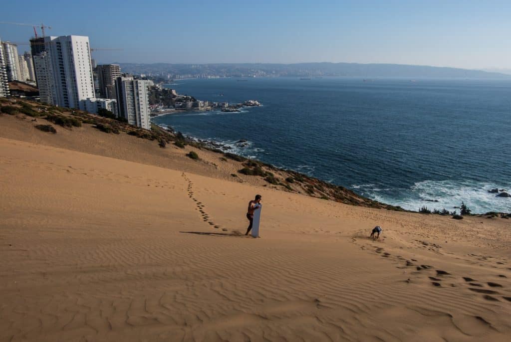 A man standing on a sand dune holding a surfboard, tall skyscrapers and the ocean in the background.