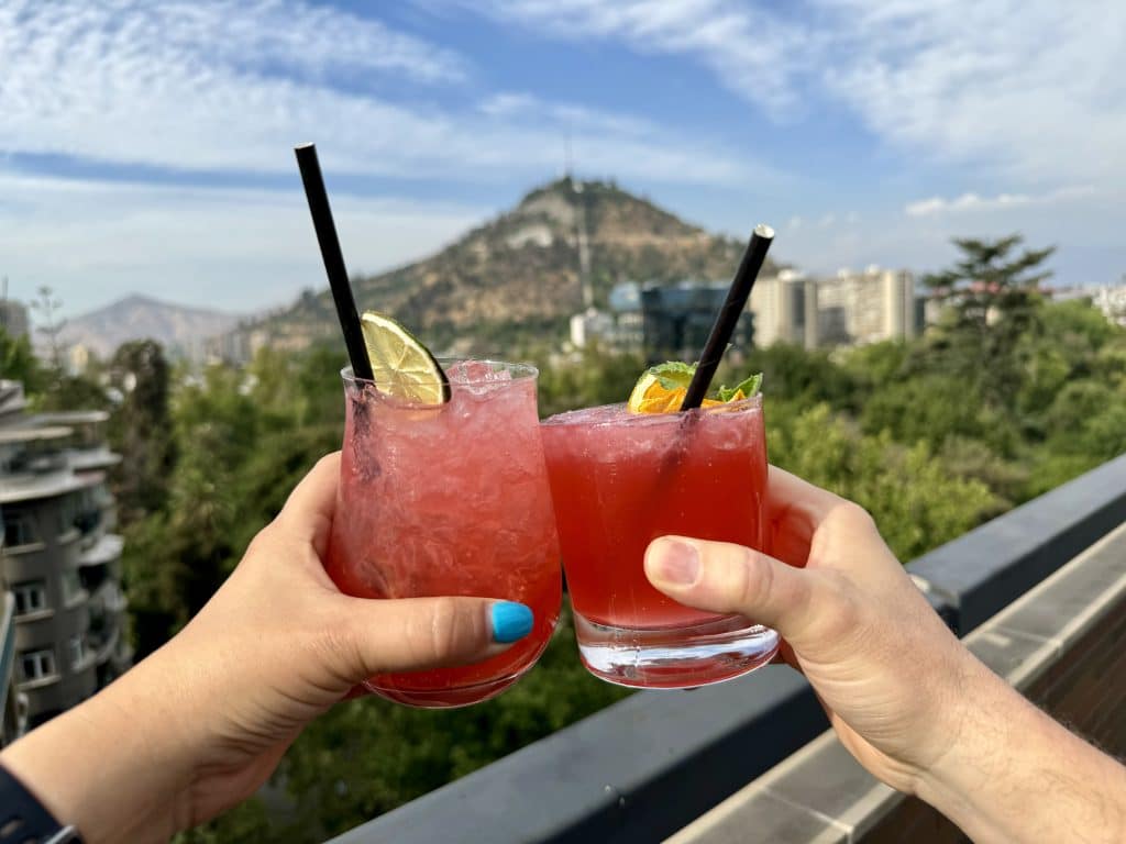 Two hands toasting red cocktails in Santiago, a mountain in the background.