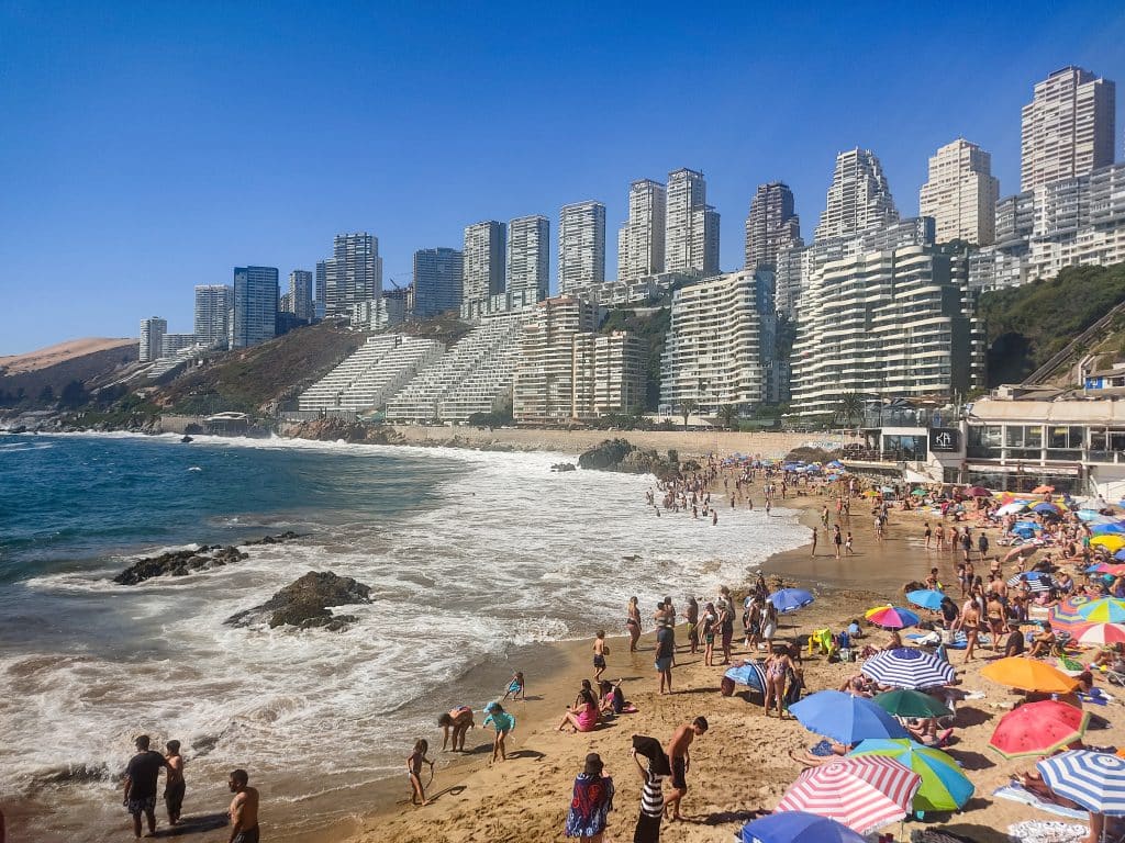 A packed, busy beach with crashing waves, and lots of tall skyscrapers in the background.