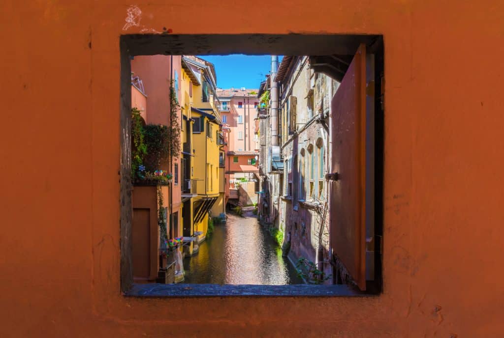 A window leading to a small, shallow canal in Bologna, warm-colored buildings on each side.