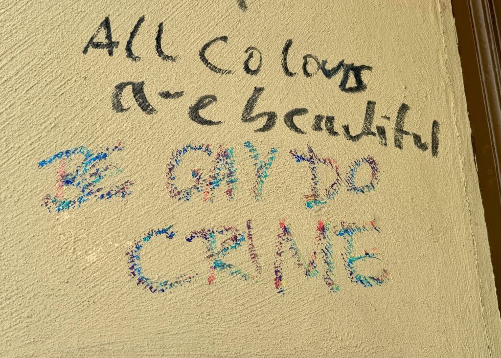 Graffiti on a wall. In black it reads "All colors are beautiful." Beneath that in rainbow crayon it reads "BE GAY DO CRIME."